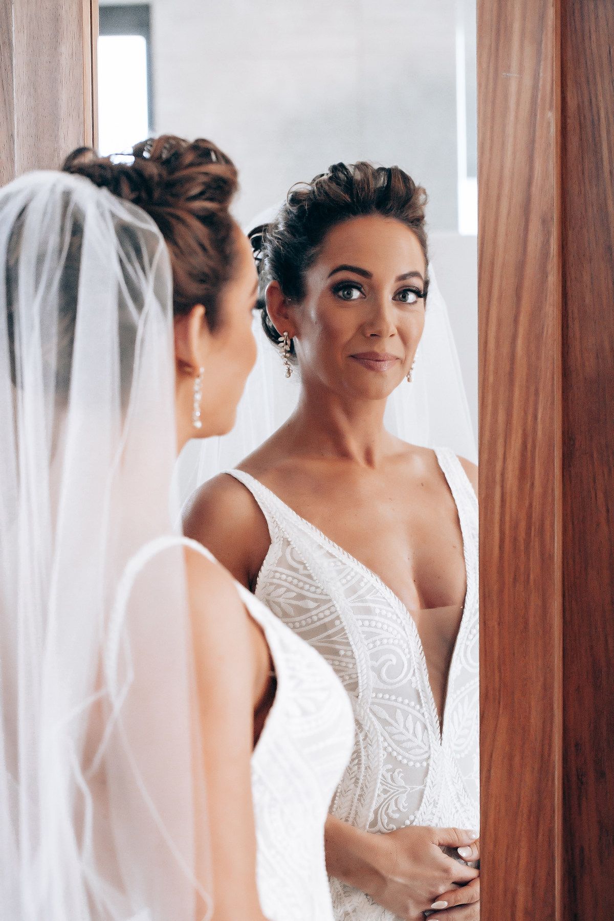 Elegant bride in lace wedding dress looking at her reflection in wooden-framed mirrors.