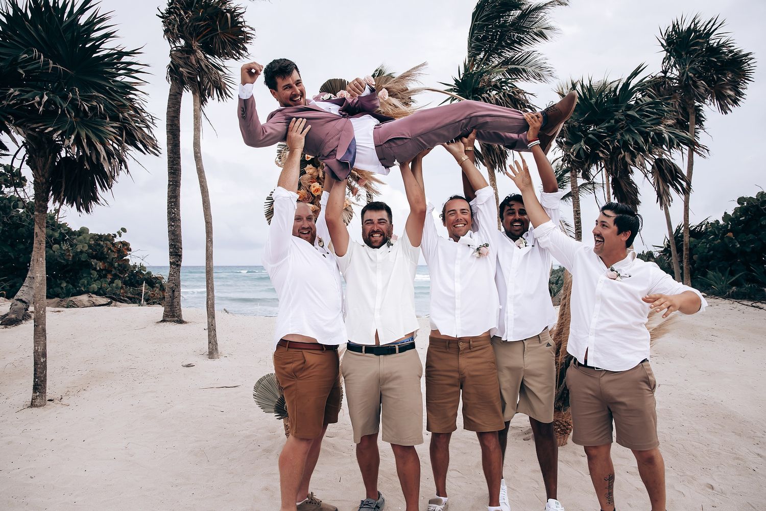 Group celebrating on a beach with palm trees, wearing coordinated white and khaki attire.