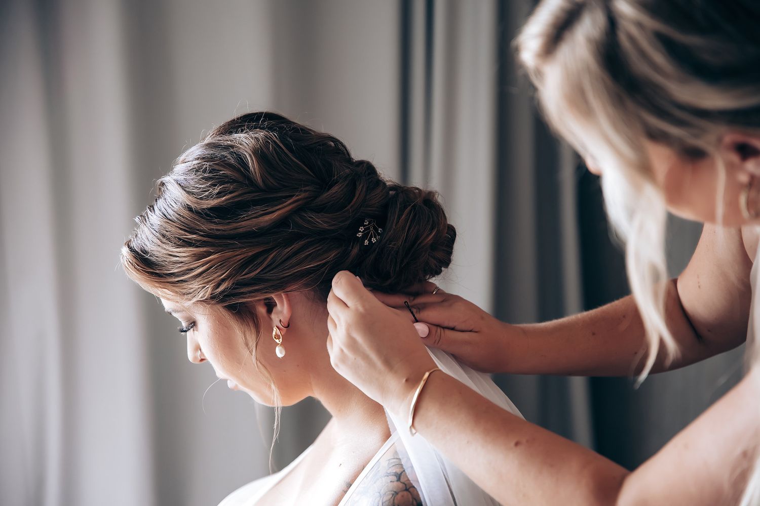 A hairstylist arranges a bride's elegant updo hairstyle while preparing for a wedding ceremony.