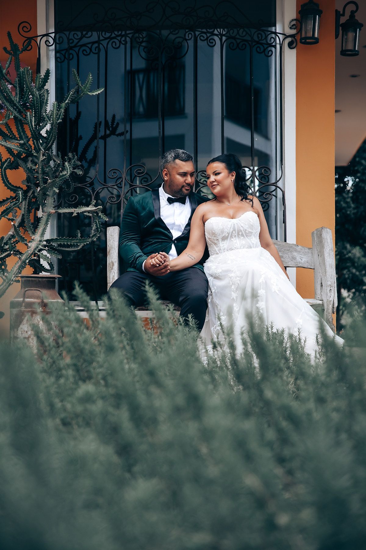 A romantic wedding couple poses together on a porch with decorative iron railings and yellow walls in the background.