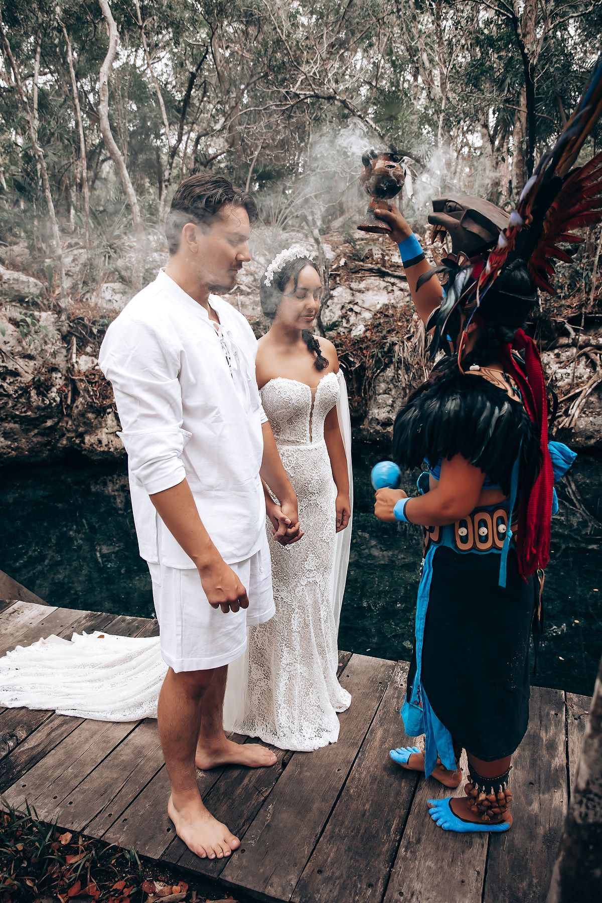 A couple in white attire stands on a wooden deck surrounded by lush tropical foliage during an outdoor ceremony.