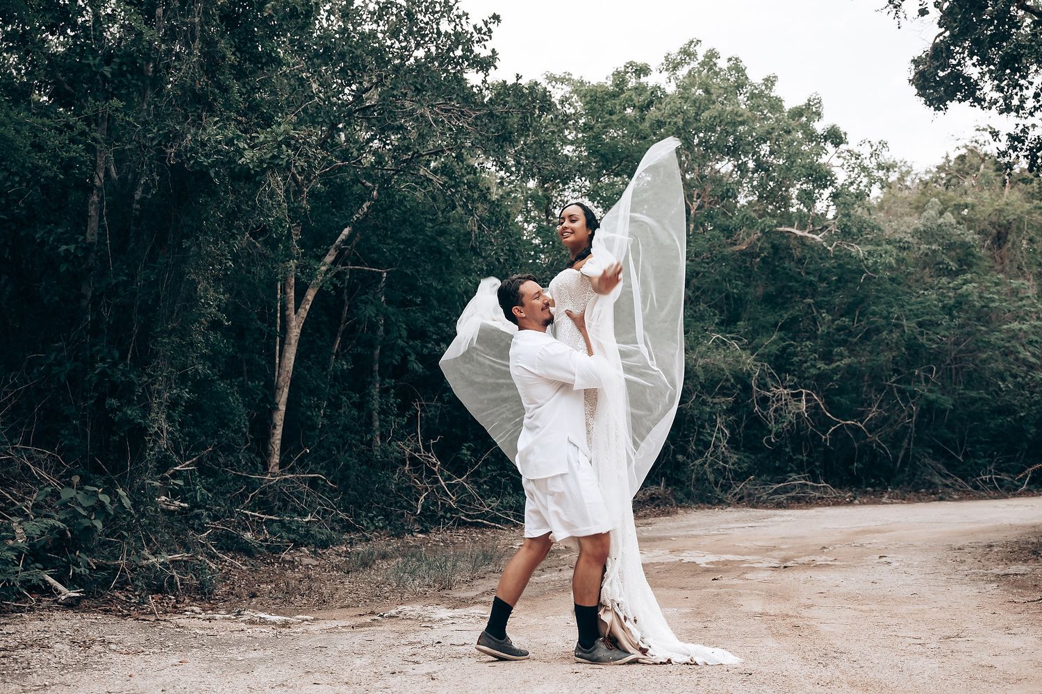 A romantic outdoor photo shoot featuring a couple in white outfits dancing and embracing on a dirt path.