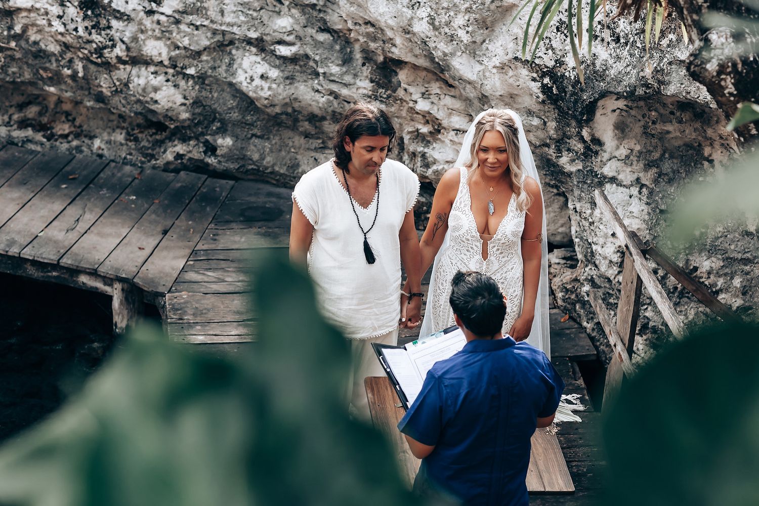 A couple walks along a scenic wooden boardwalk near a rocky cliff and turquoise water on a sunny day.