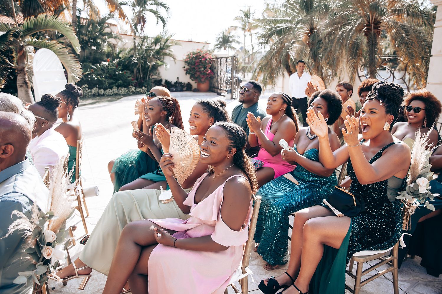 Wedding guests celebrate and cheer during an outdoor ceremony surrounded by palm trees.