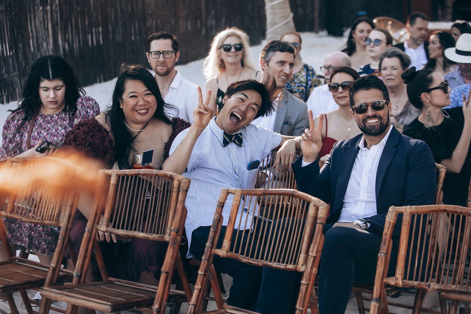 Wedding guests seated on wooden benches laugh and smile during an outdoor ceremony.