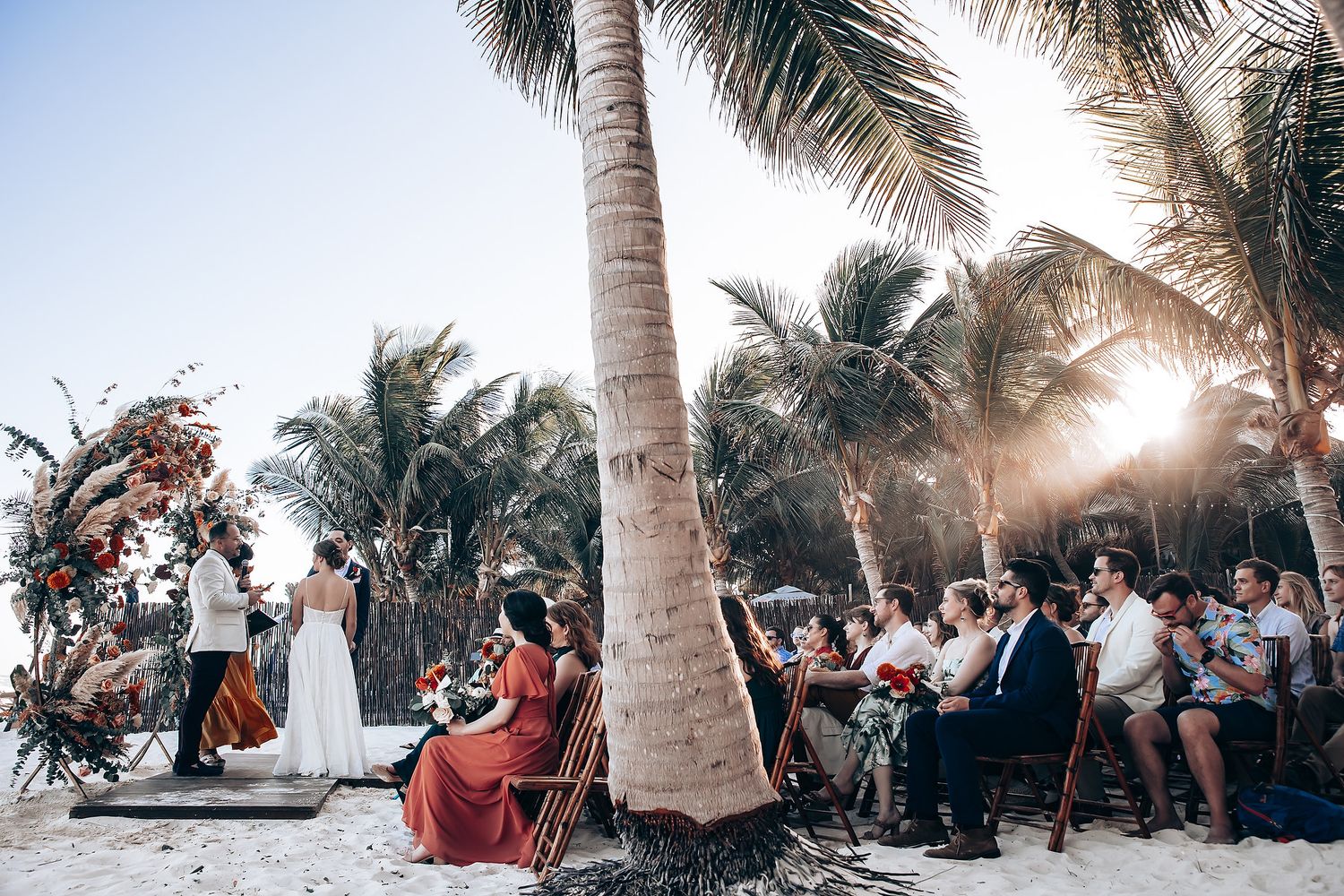 A beachside wedding ceremony under palm trees at sunset with guests seated on wooden chairs.