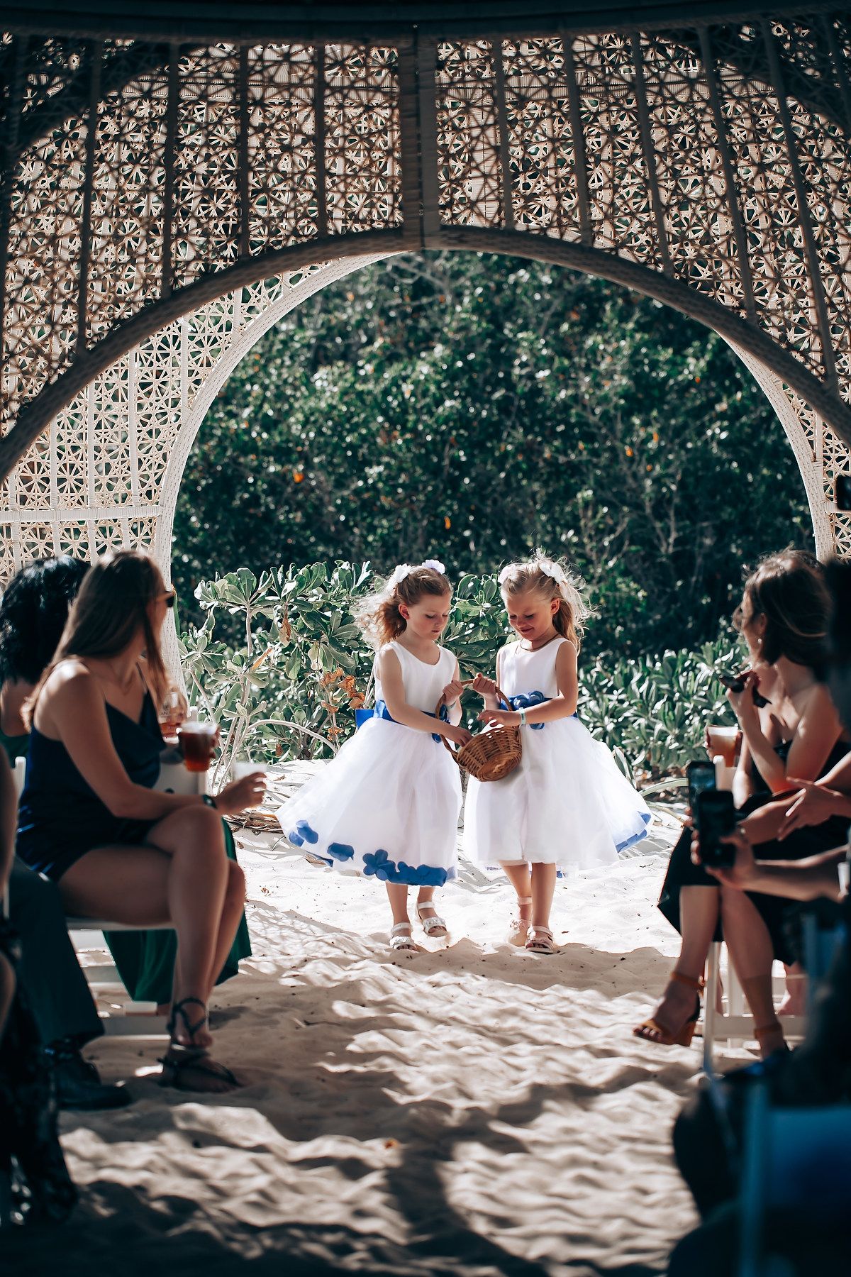 Children in white dresses playing together on sandy ground under a decorative latticed gazebo structure.