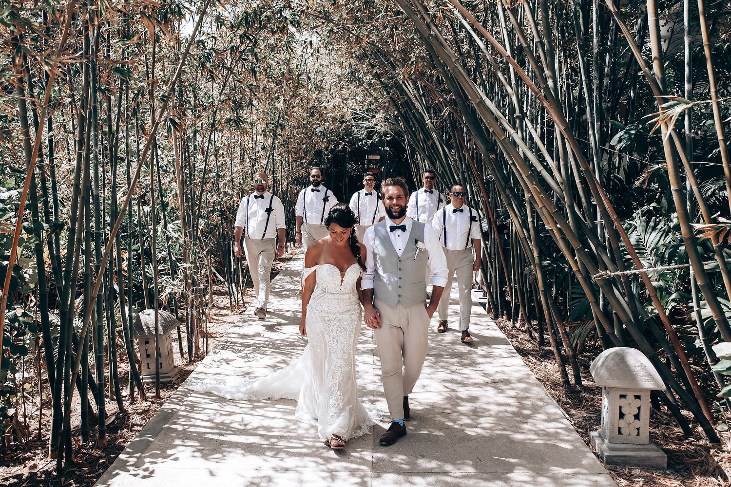Wedding party members walk through a tree-lined pathway in coordinated white and beige attire.