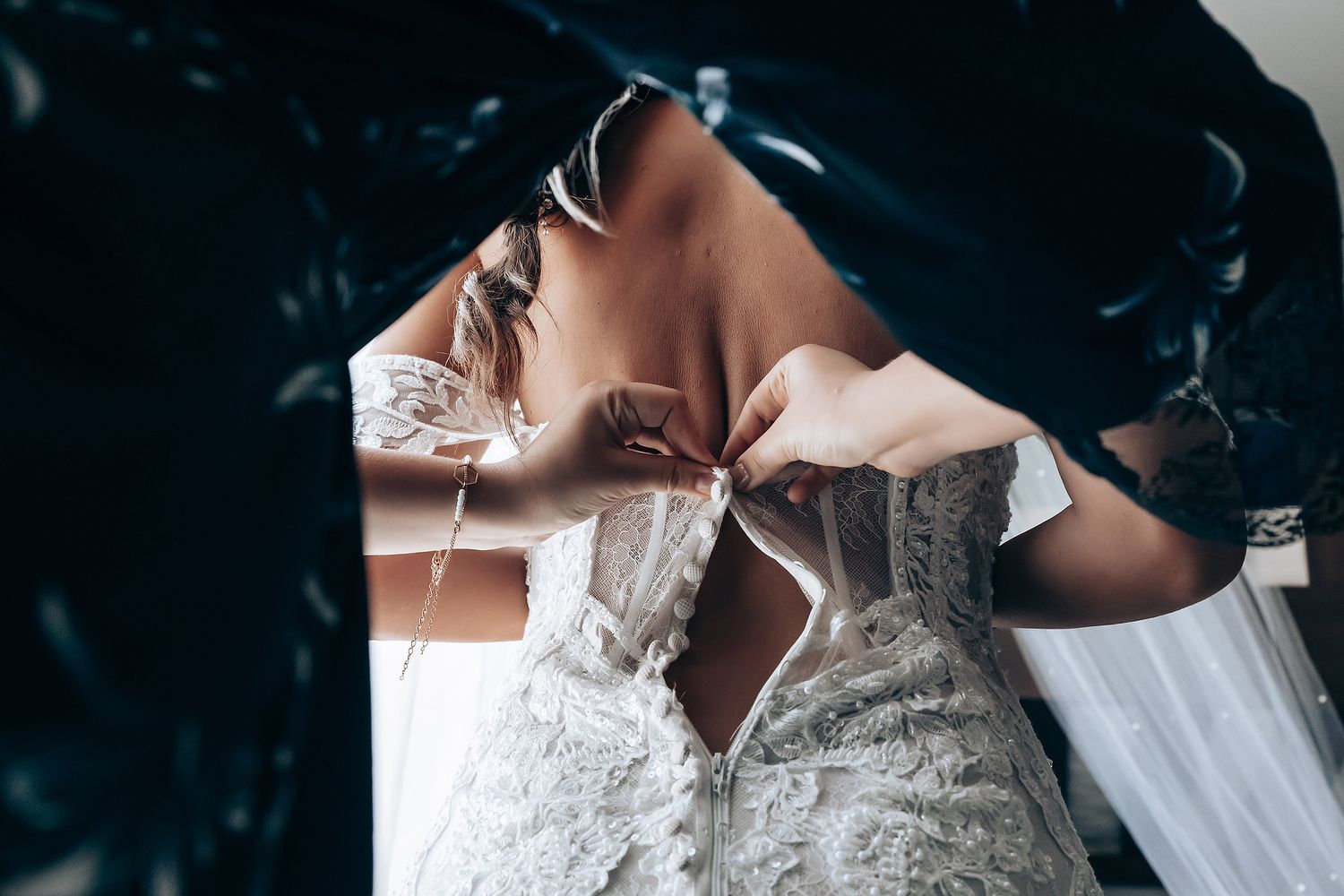 Someone lacing up a white lace wedding dress with intricate beaded details against a dark background.