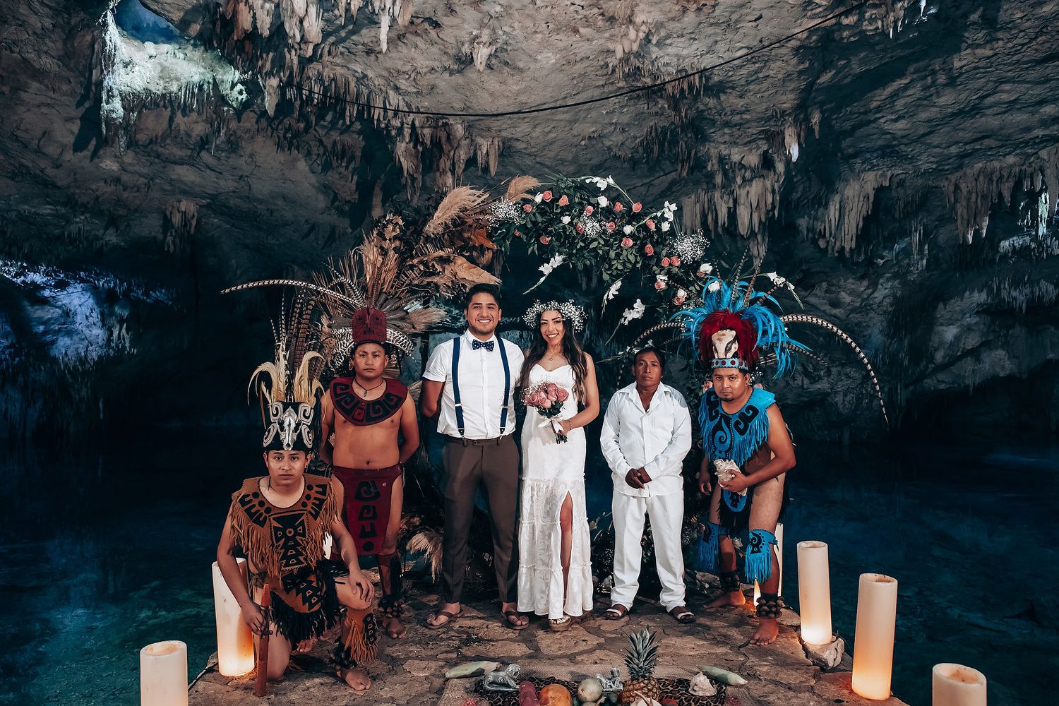 A group wedding photo showing attendees in white and colorful outfits gathered inside a dimly lit cave environment.