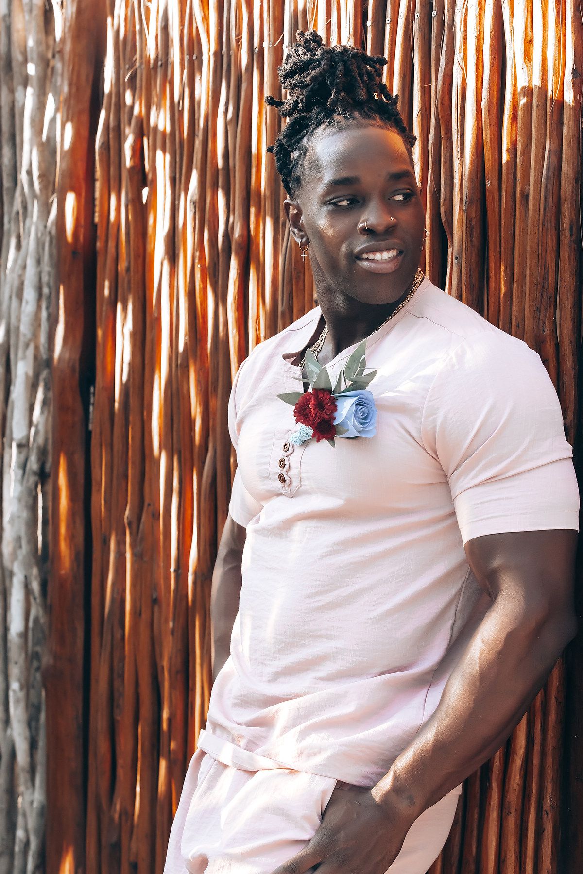 A smiling young Black man wearing a pink shirt and standing in front of a wooden wall.