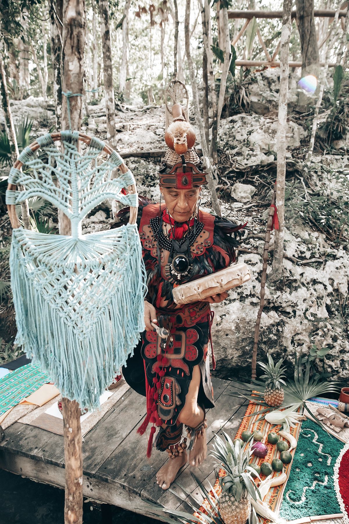 A person dressed in traditional indigenous attire standing on a wooden platform in a forested setting.