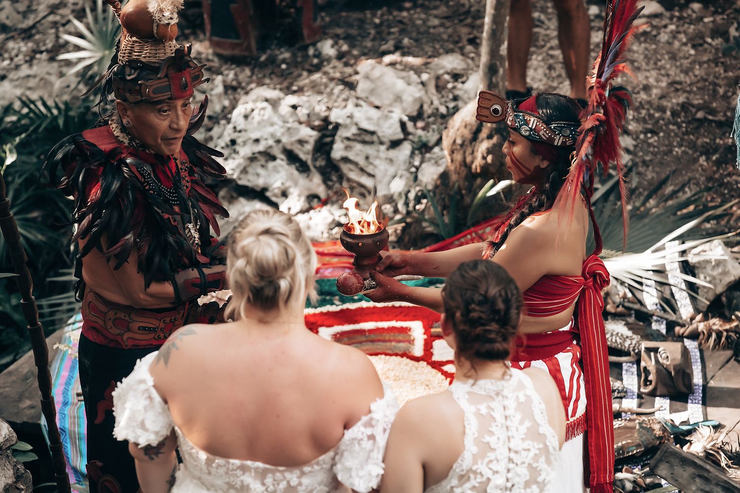 Two brides in white wedding dresses standing together with a person in traditional indigenous attire.