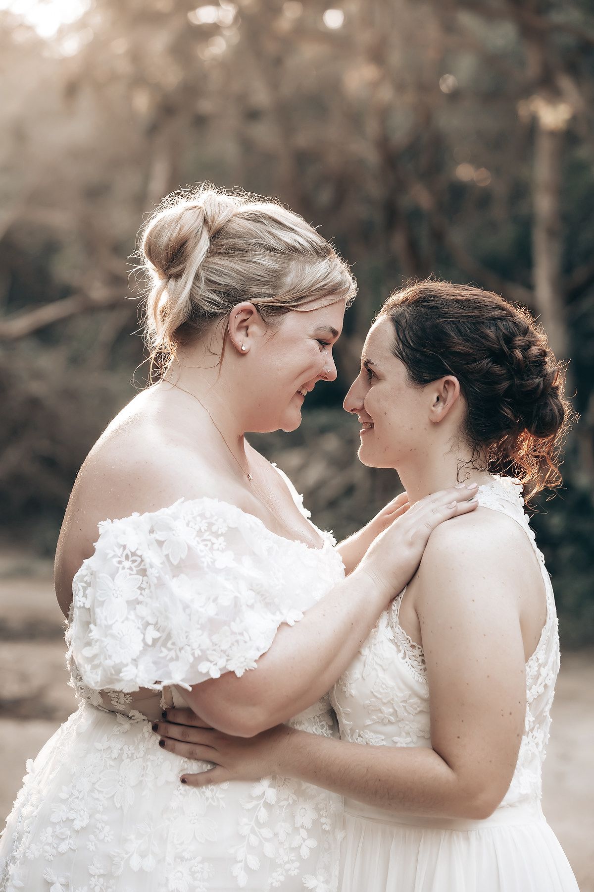 Two brides in white wedding dresses embrace each other lovingly in a natural outdoor setting.