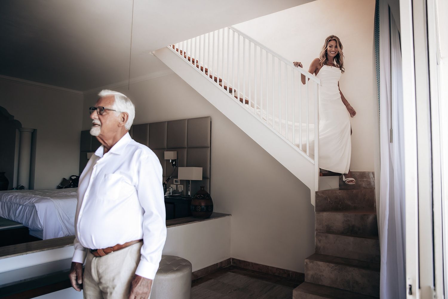 An older man stands in a room, while a woman in a white dress walks up the stairs.