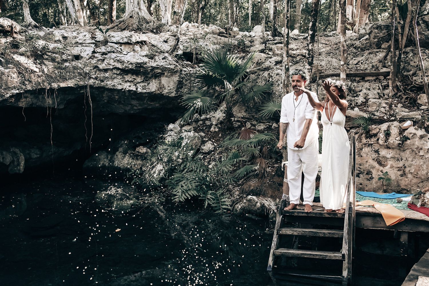 A couple in white clothing stands on a wooden bridge over a natural pool in a forest.