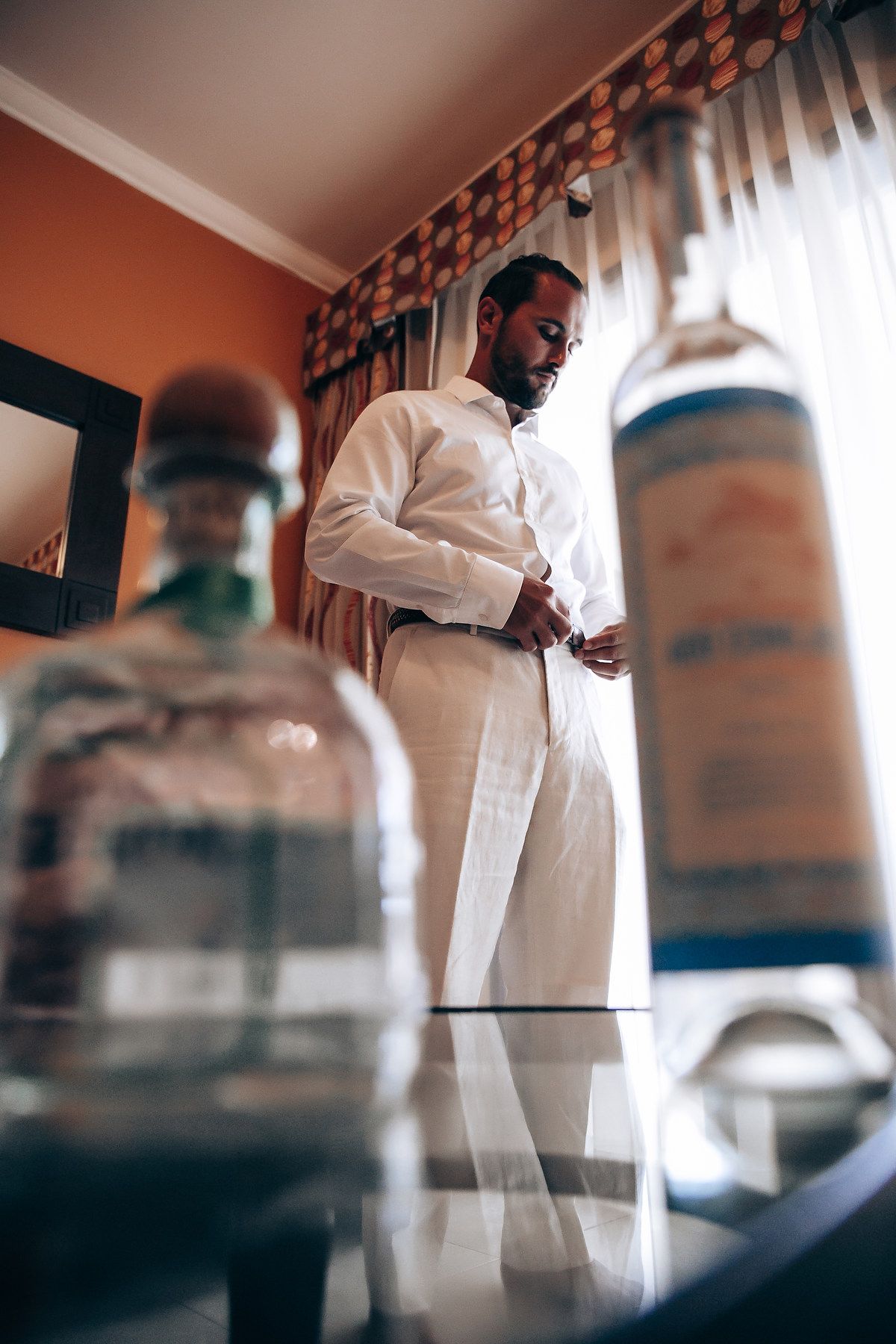 A person in white attire stands near glass bottles in a room with orange walls and curtains.