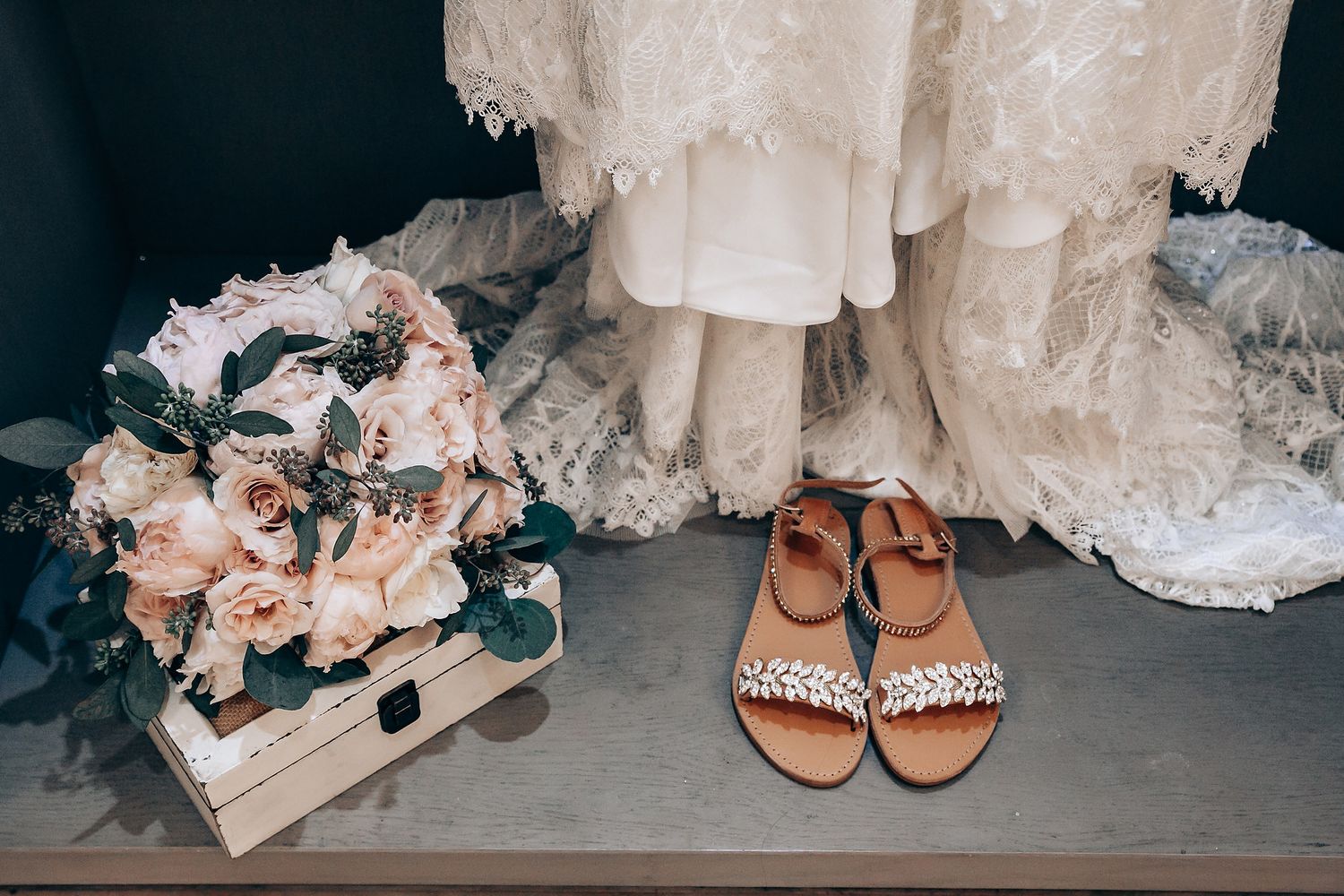 Wedding accessories including a floral bouquet and jeweled sandals displayed next to a lace dress.
