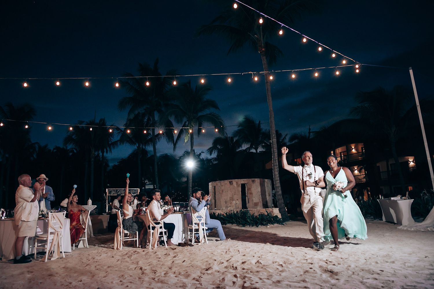 String lights illuminate nighttime beach wedding reception with guests dancing on sand.