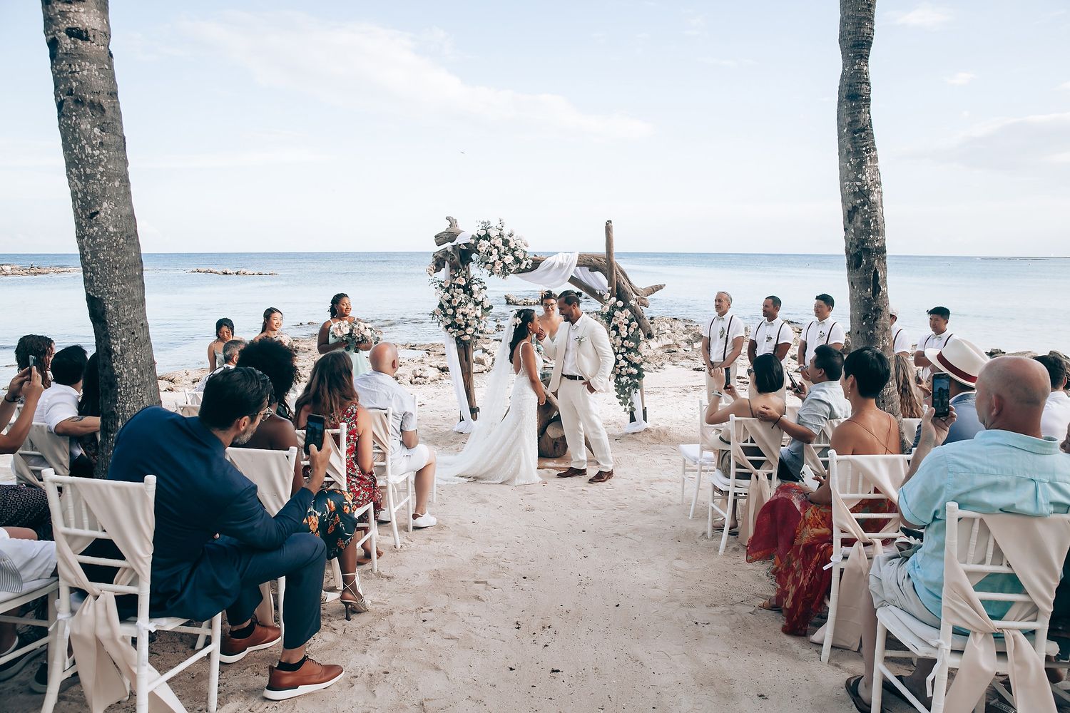 Beachside wedding ceremony with guests seated in white chairs between palm trees.