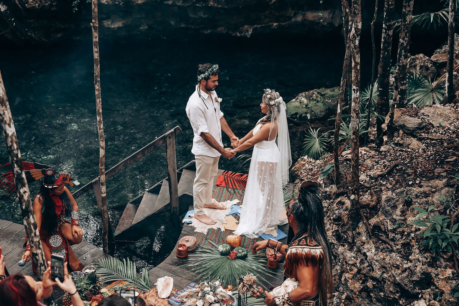 An intimate outdoor wedding ceremony surrounded by tropical foliage and decorative elements near a dark cave.