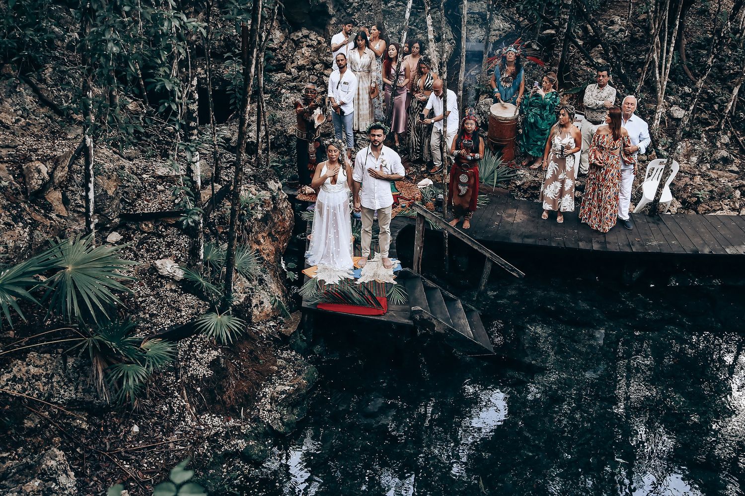 Wedding ceremony participants gathered along a natural cenote or water feature in tropical forest setting.