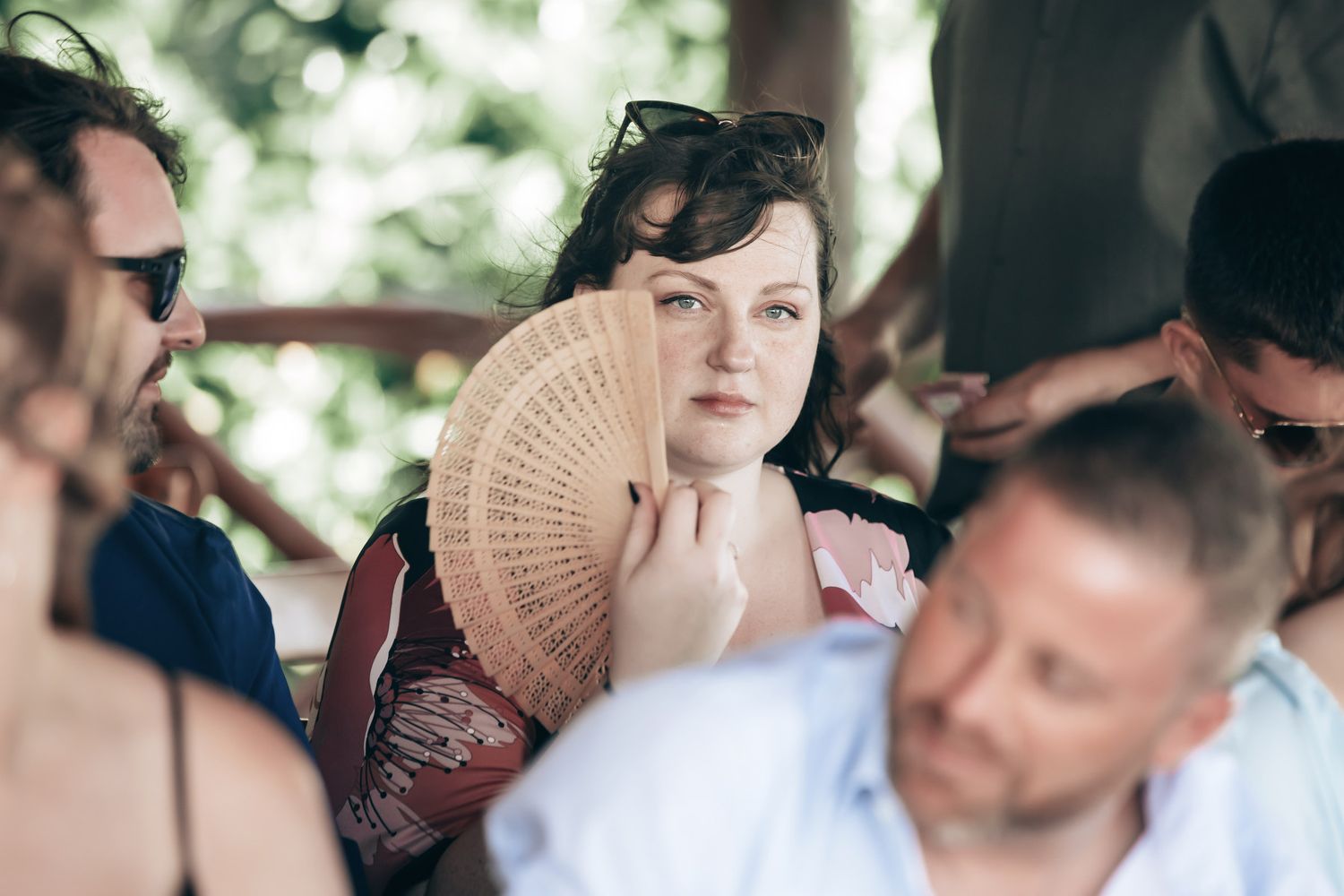 Wedding guest holding a wooden hand fan while seated among other guests at an outdoor ceremony.