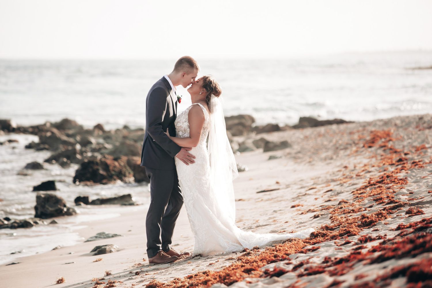 Newlyweds share a romantic kiss on a scenic beach with ocean waves and rocky shoreline at sunset.