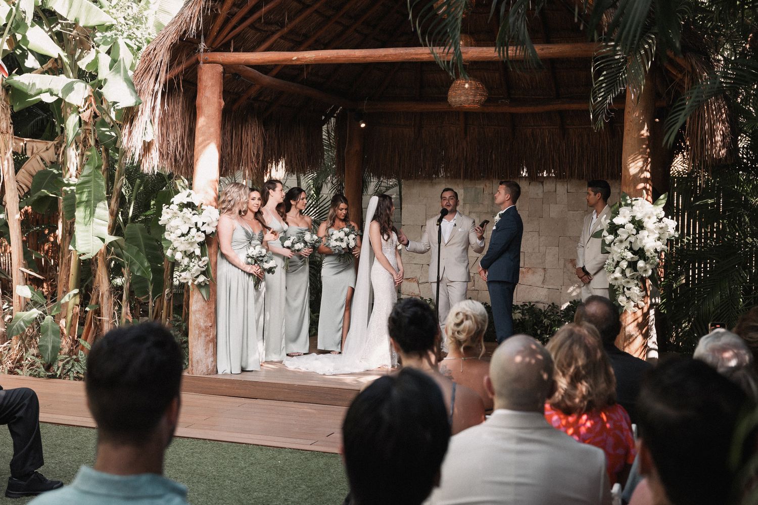 Wedding ceremony under rustic thatched roof altar with bridesmaids in sage green and floral arrangements.