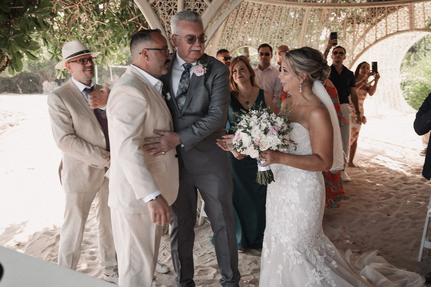 Bride in lace gown and groom in cream suit greeting guests during outdoor beach wedding ceremony.