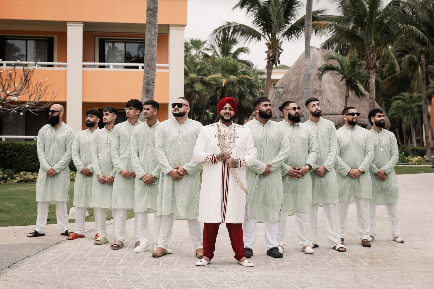 Sikh groom in white sherwani and red turban poses with groomsmen in matching sage green kurtas at outdoor wedding.