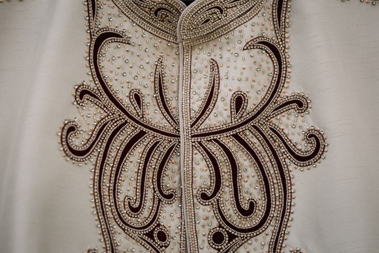Close-up of intricate dark brown embroidery on white fabric featuring elegant scrollwork and beaded detailing.