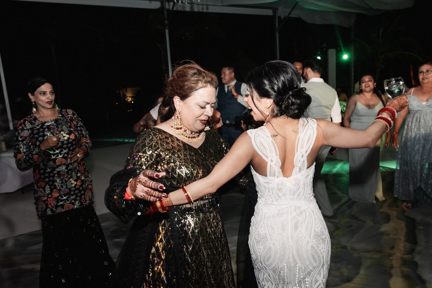 Bride in lace gown dancing with older woman in gold sequin dress at tropical wedding reception.