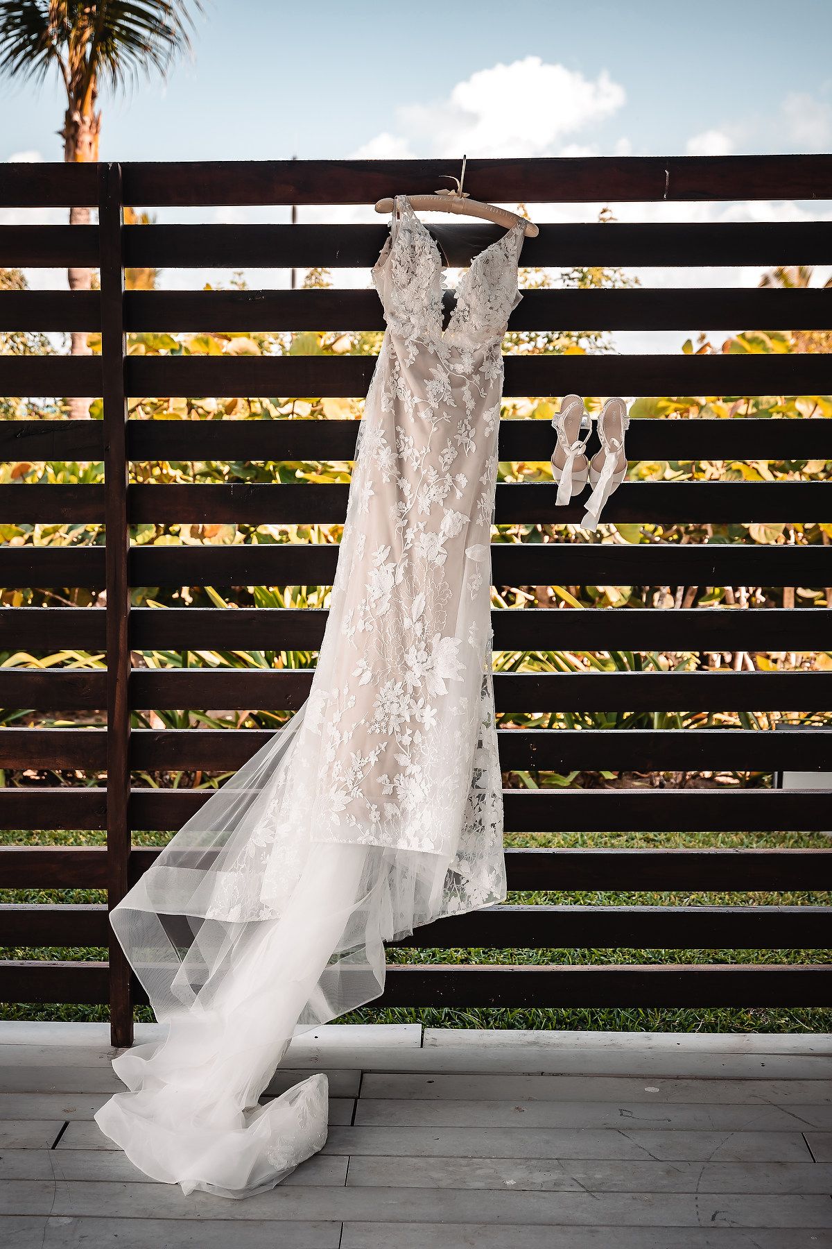 Elegant lace wedding dress with long train hanging on wooden fence with bridal heels outdoors.