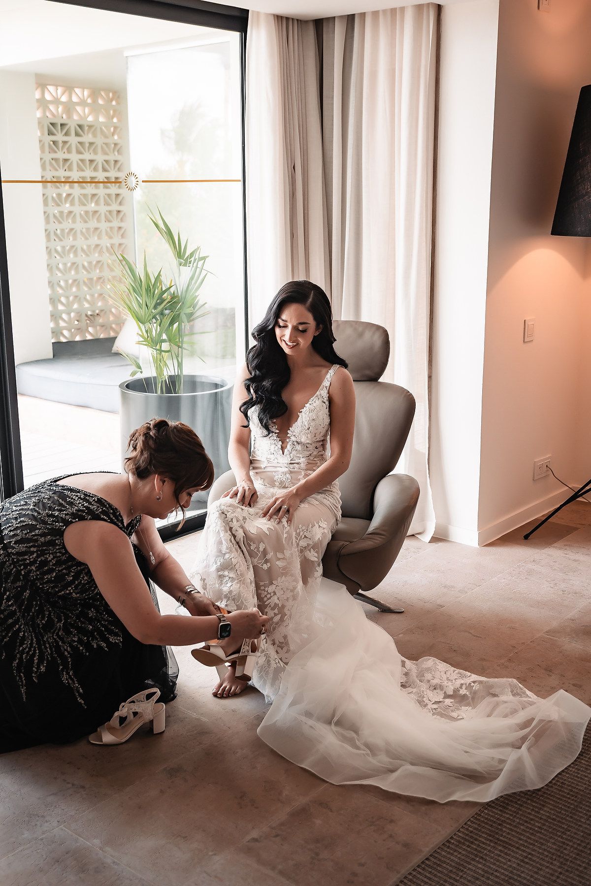 Bride in lace wedding gown seated in chair while bridesmaid helps with shoes in elegant hotel suite.