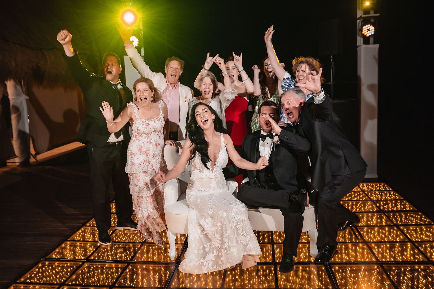 Joyful wedding party celebrating on a glowing dance floor at night, with bride in white lace gown surrounded by cheering guests.