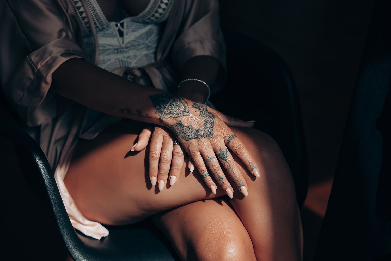 Close-up shot of tattooed hands resting on legs in a dark moody setting.