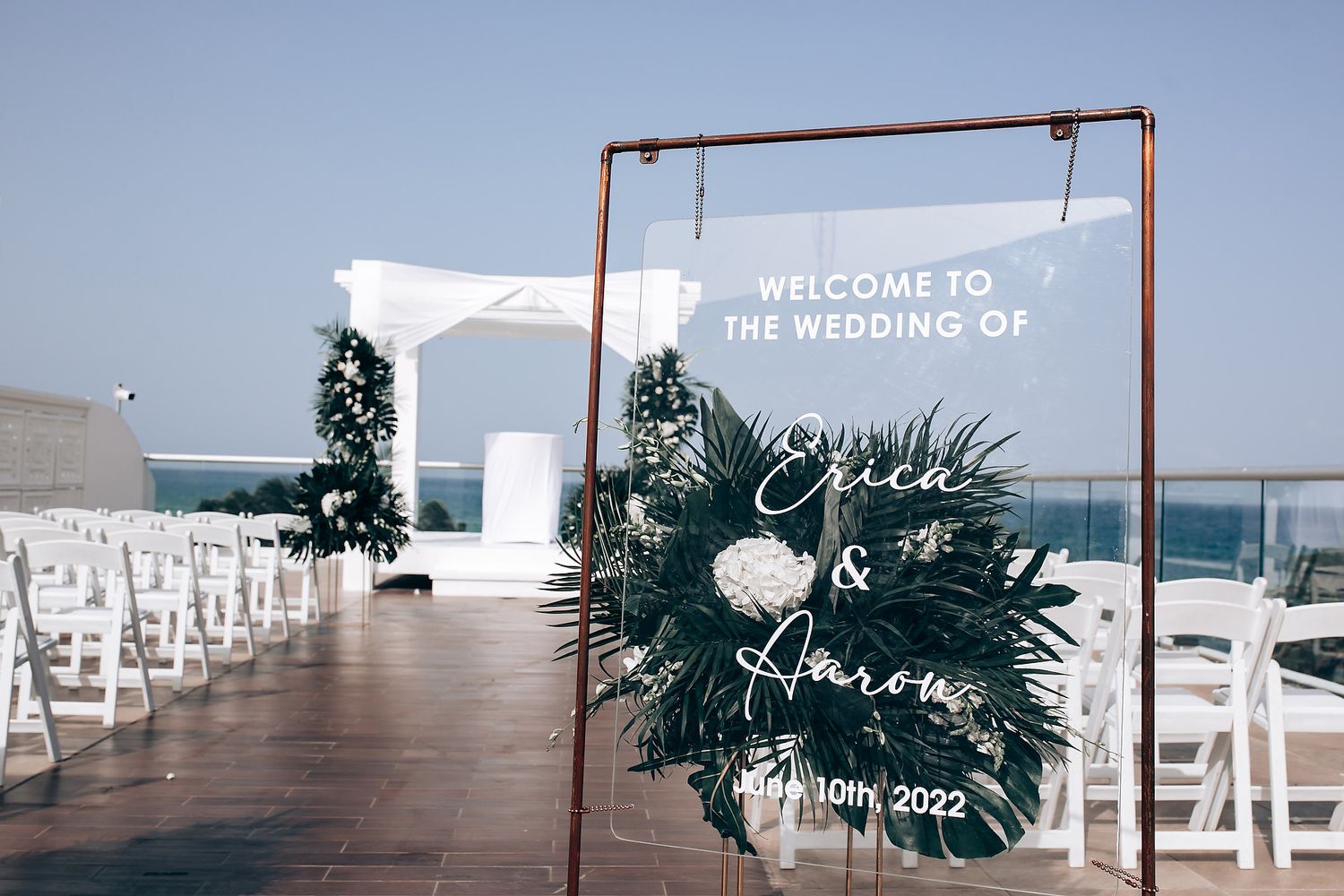 Modern copper wedding arch with greenery and white chairs overlooking the ocean.