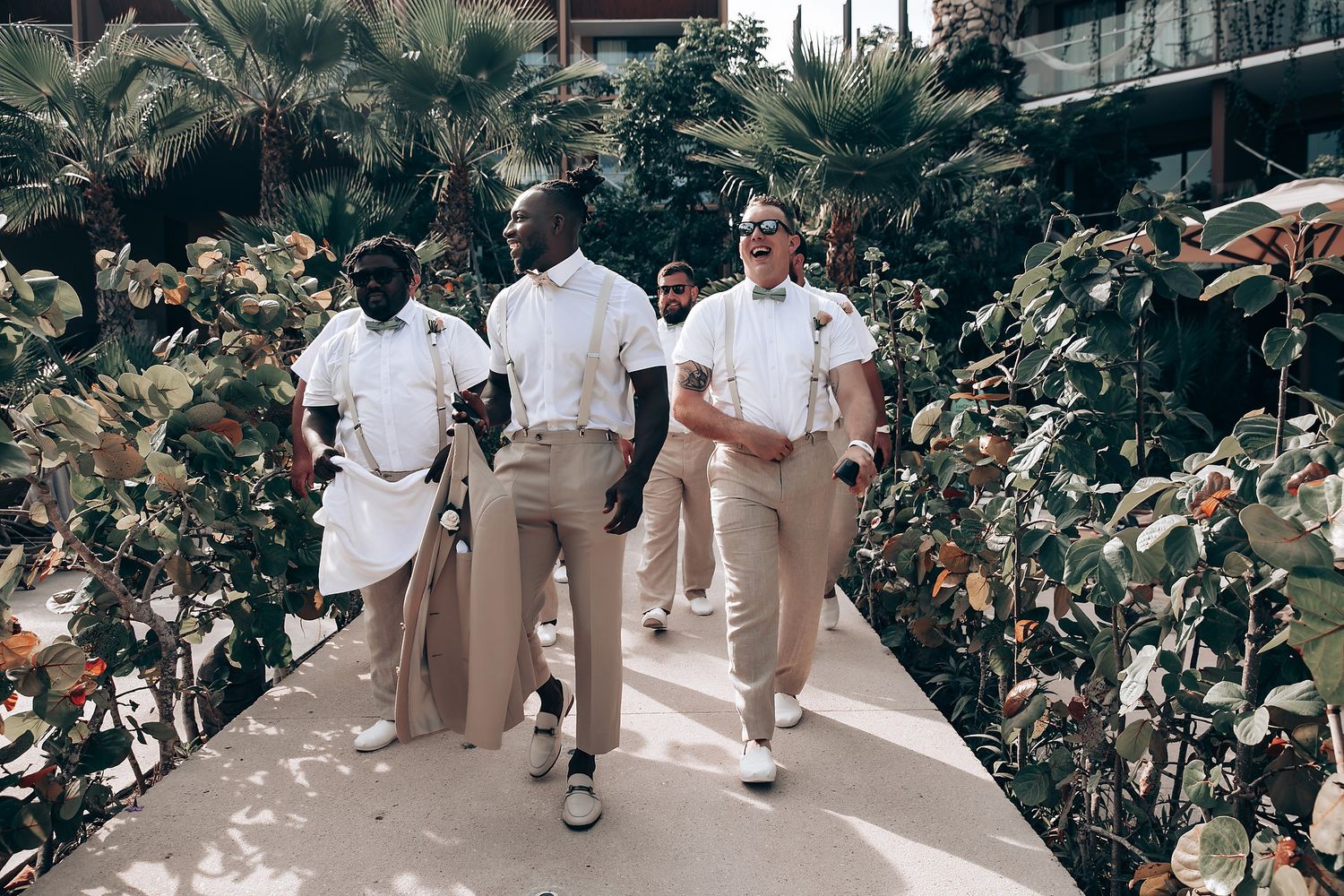 Wedding party members walk through a lush garden pathway wearing coordinated neutral-toned outfits.