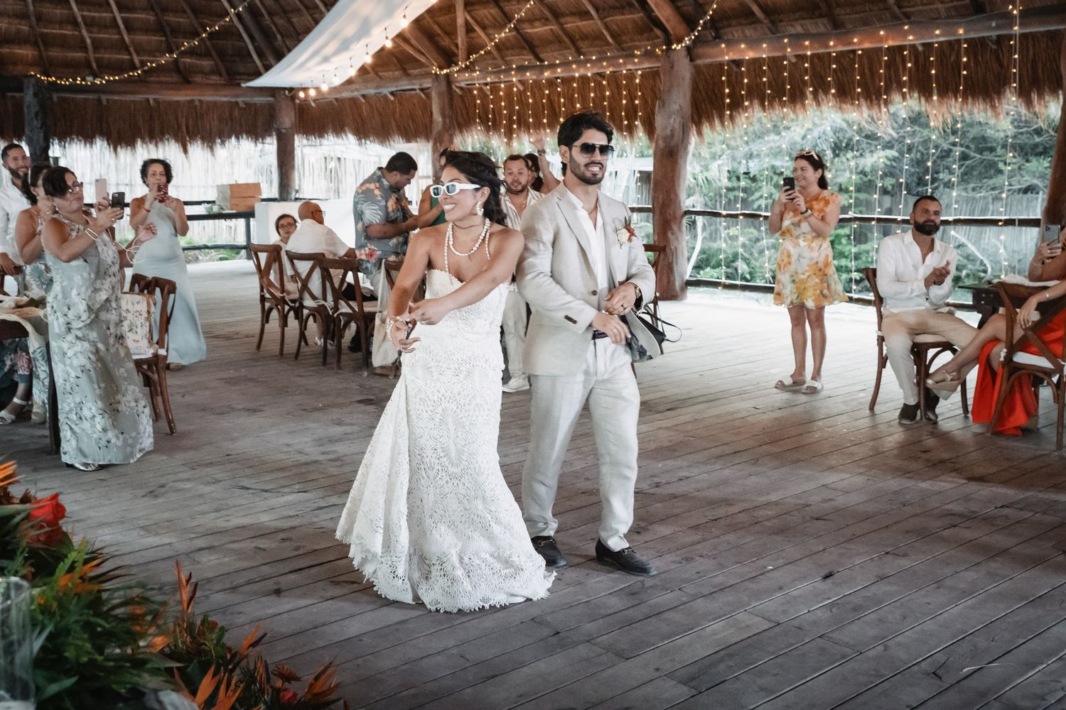 Bride and groom enter reception at tropical outdoor venue with string lights and floral decor.