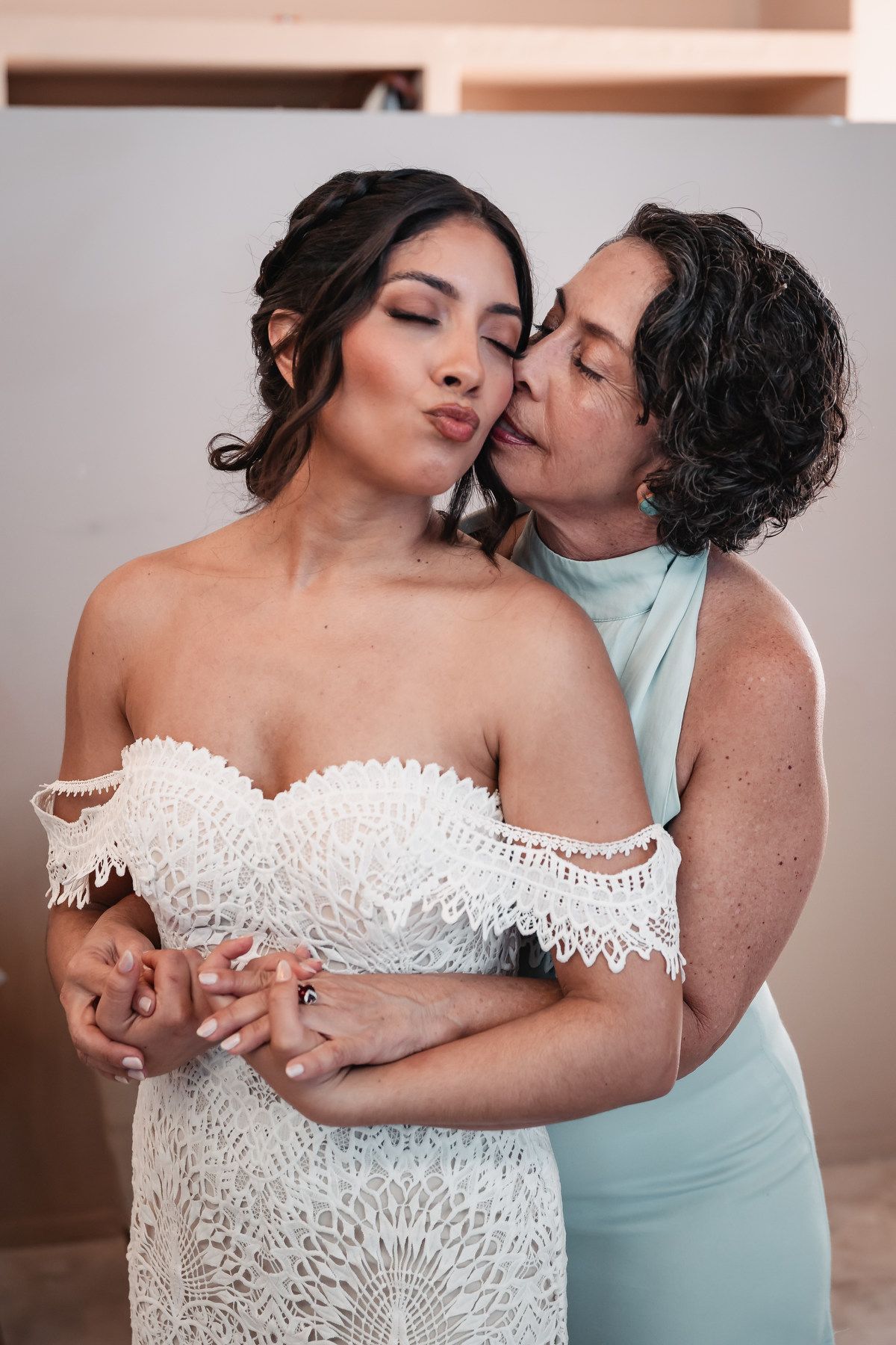 Older woman in mint dress embracing and kissing bride in off-shoulder lace wedding gown.