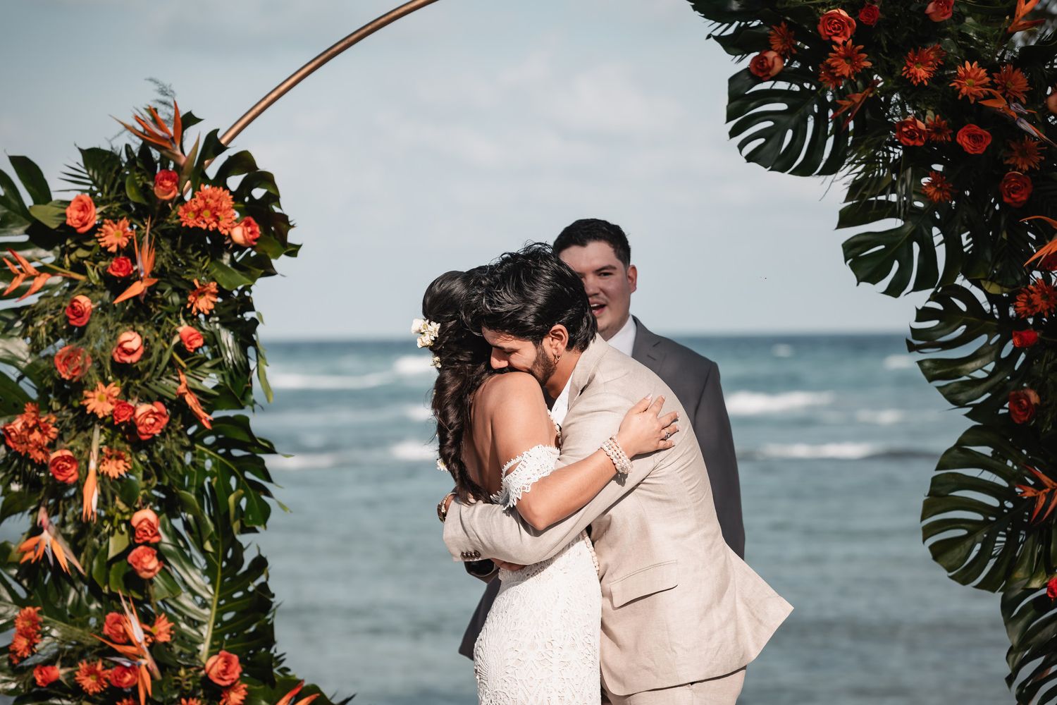Bride and groom embracing under tropical floral arch with ocean backdrop.