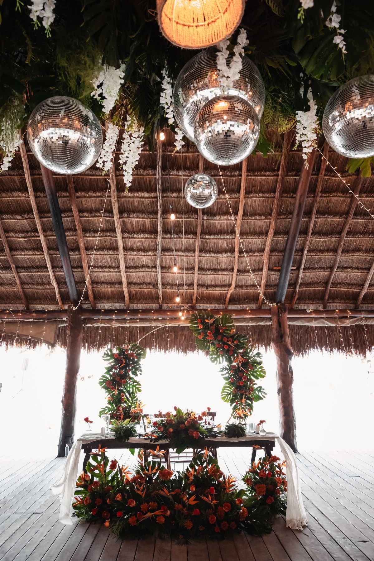 Tropical wedding reception table adorned with disco balls, lush greenery, and vibrant floral arrangements under a thatched roof.
