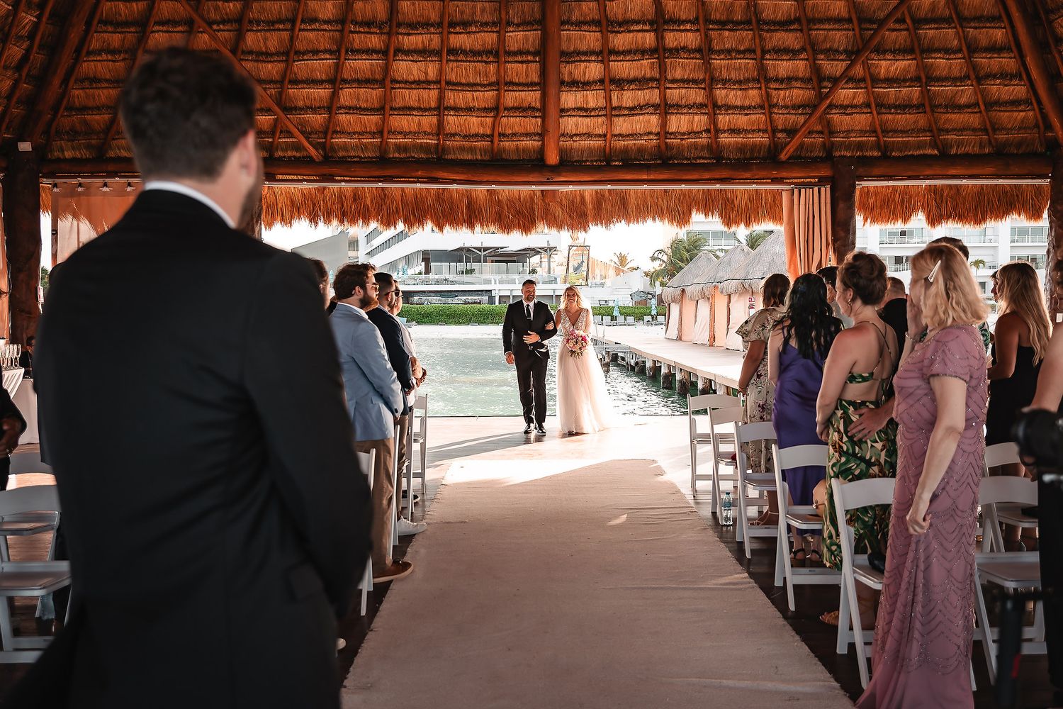 Wedding ceremony setup under rustic thatched roof palapa with white chairs.