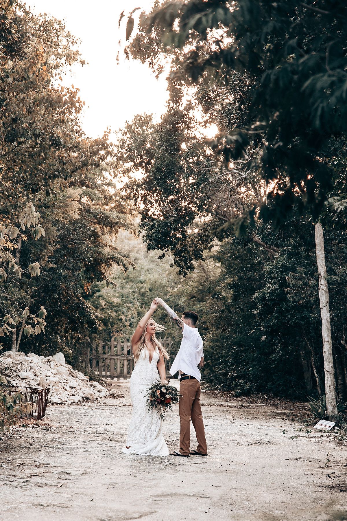 Romantic couple in white dress and brown attire walking together on a dirt path surrounded by lush trees.
