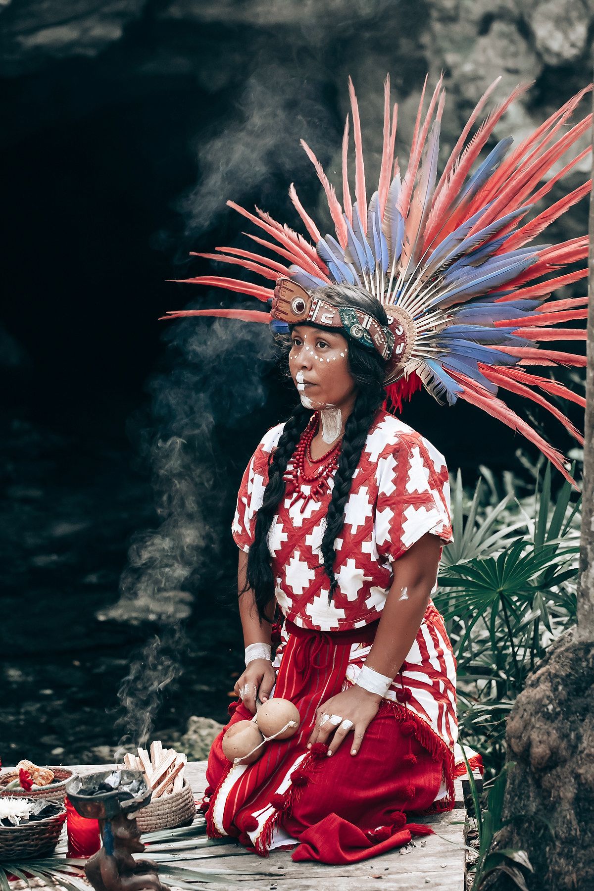 Performer wearing red and white traditional dress with colorful feathered headdress sits in natural setting.