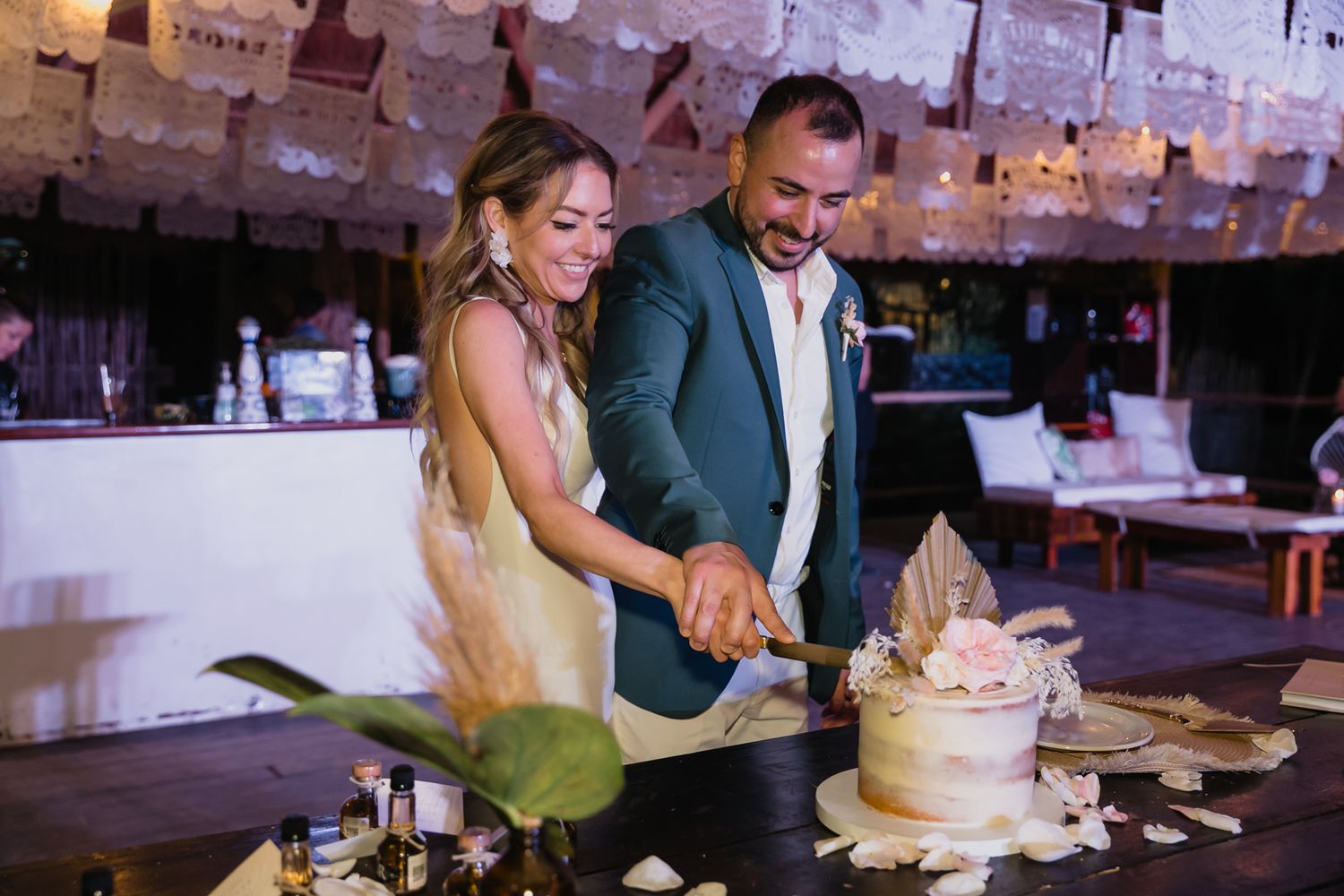 Smiling bride and groom cut wedding cake together at tropical reception venue.