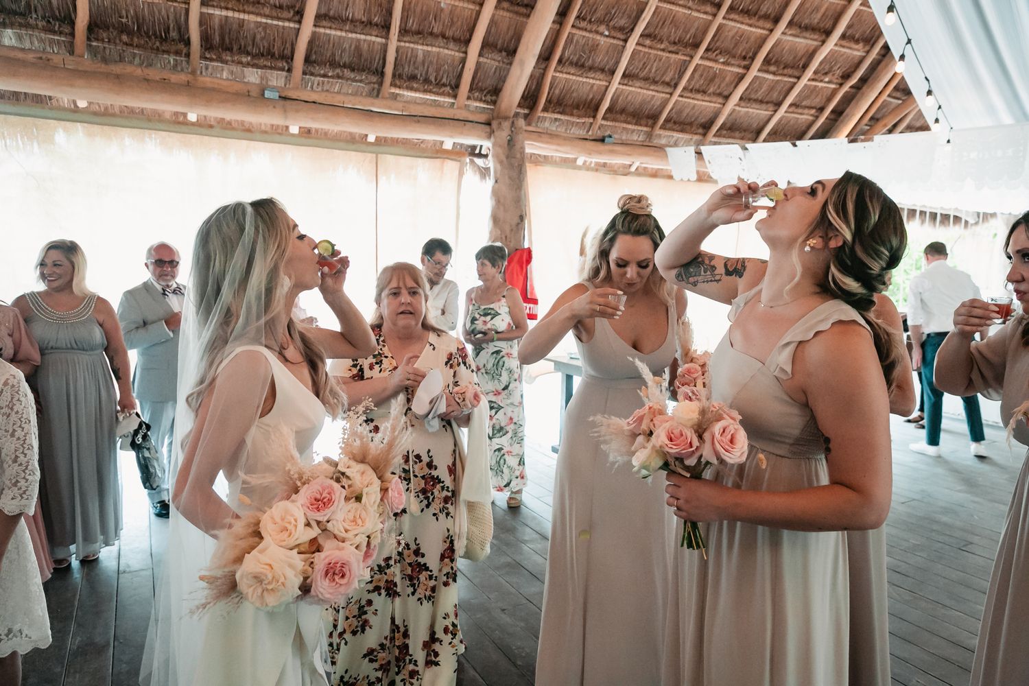 Bride and bridesmaids take celebratory shots while holding pink bouquets at tropical beach wedding reception.