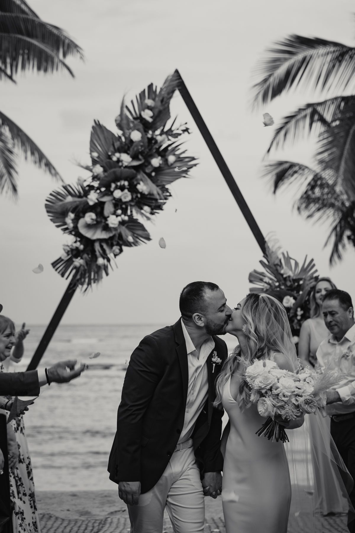 Black and white photo of newlyweds sharing first kiss under floral arch on tropical beach as guests celebrate.