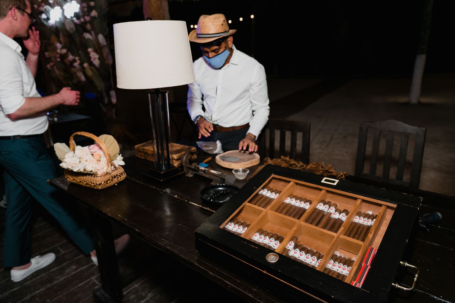 Cigar roller in straw hat preparing cigars at a wooden display table during a wedding reception.